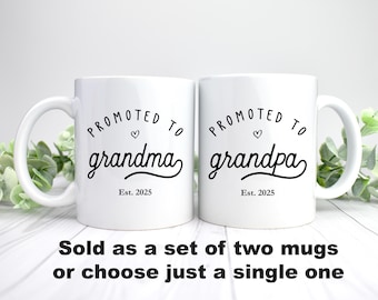 May include: Two white ceramic mugs with black text. The mug on the left says "PROMOTED TO grandma Est. 2025" with a small heart symbol. The mug on the right says "PROMOTED TO grandpa Est. 2025" with a small heart symbol.