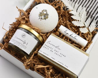 Könnte beinhalten: Eine weiße Geschenkbox gefüllt mit braunem, zerkleinertem Papier. Die Box enthält eine weiße Badebombe, ein kleines Glas mit goldenem Deckel, das mit "(serenity) hand-poured candle" beschriftet ist, eine Seife mit der Aufschrift "lavender beauty bar soap pain de savon all natural vegan cruelty free" und eine kleine Tube Lippenbalsam mit der Aufschrift "lip balm".