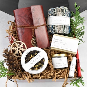 May include: A gift box containing a red leather journal, cosy socks, lavender soap, a candle, lip balm, and loose leaf tea. The journal features a leaf design and a metal leaf charm. The box is lined with brown paper and a wooden snowflake ornament.