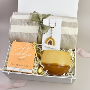 May include: A white gift box filled with a beige and white striped throw blanket, a yellow and brown mug, a gold spoon, a box of "Cup of Sunshine" herbal tea, and a rainbow-coloured keychain.