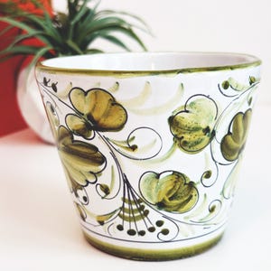 May include: White ceramic planter with a hand-painted floral design in shades of green and olive. The planter has a green rim and base, and the floral pattern includes stylized leaves and flowers. A decorative item for indoor plants.