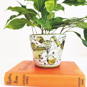 May include: A potted plant with vibrant green leaves sits atop an orange book. The ceramic pot features a white base with a green and yellow floral design. The book is titled "Mystery House" by Kathleen Norris.