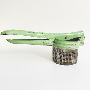 May include: A vintage green metal potato masher with a round, perforated base. The masher has a long handle with a slight curve.