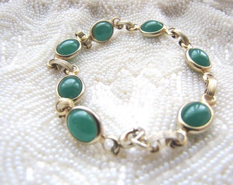 May include: A gold-toned bracelet featuring oval, emerald-green stones. The stones are set in a gold bezel and connected by gold links. The bracelet is displayed on a white, textured surface, creating a contrast between the jewelry and the background.