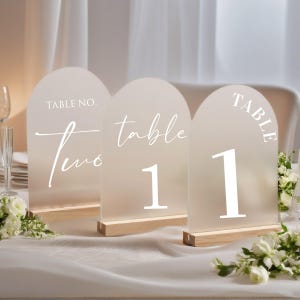 May include: Three frosted acrylic table number signs with arched tops and wooden bases. The signs display white text: "TABLE NO. Two", "table 1", and "TABLE 1". The signs are set on a white tablecloth with floral arrangements.