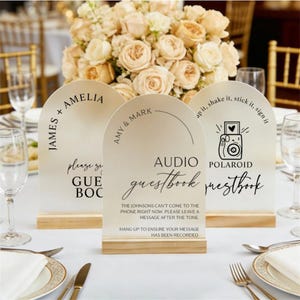 Puede incluir: Señales de mesa de boda con tapas arqueadas y bases de madera. Los letreros tienen texto que dice "James + Amelia", "Amy & Mark" y "Polaroid Guestbook". Los letreros son de color beige con texto negro.