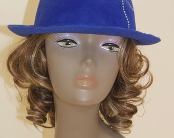 May include: A royal blue fedora-style hat with a slightly curved brim. The hat features a decorative silver-colored embellishment on the side. The hat is displayed on a mannequin head with brown and blonde curly hair.