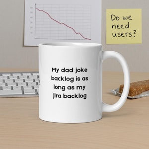 Może przedstawiać: Biały ceramiczny kubek z czarnym napisem "My dad joke backlog is as long as my Jira backlog". Kubek ma zakrzywiony uchwyt i stoi na jasnobrązowej powierzchni.