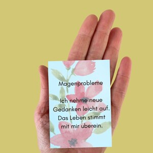May include: A small, rectangular card with text and floral illustrations is held in a hand. The card features the words "Magenprobleme", "Ich nehme neue Gedanken leicht auf. Das Leben stimmt mit mir überein."