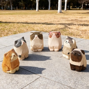 May include: A collection of six wooden cat figurines, each with unique markings and expressions. The cats are carved from light-colored wood and painted with brown, gray, and tan details. They are arranged on a gray stone surface with a park background.
