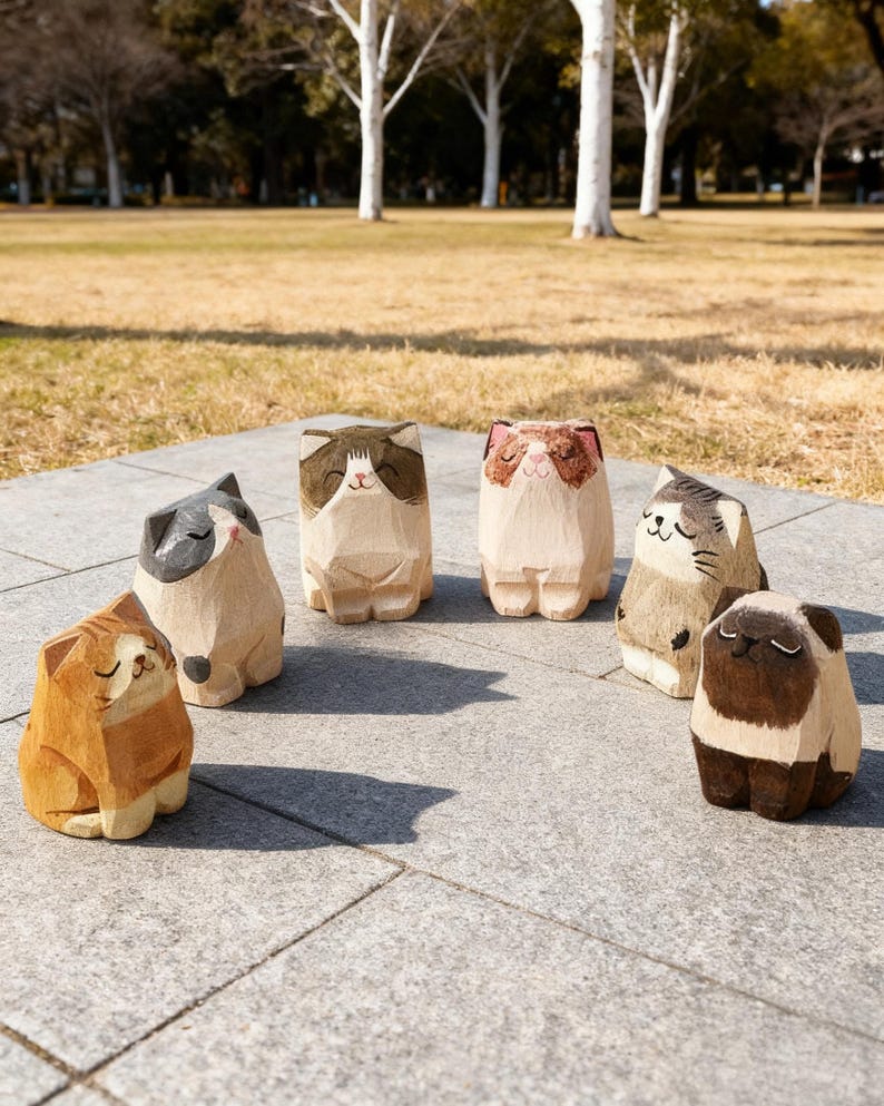 May include: A collection of six wooden cat figurines, each with unique markings and expressions. The cats are carved from light-colored wood and painted with brown, gray, and tan details. They are arranged on a gray stone surface with a park background.