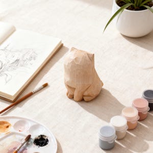 May include: A carved wooden cat figurine sits on a linen surface, next to a sketchbook, paint palette, and small paint pots. The cat is light brown, with visible carved details. A small houseplant in a white pot is in the background.