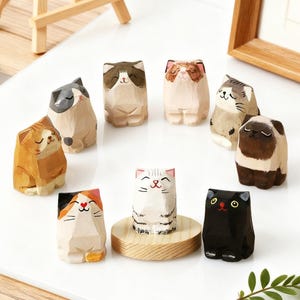 May include: A collection of hand-carved wooden cat figurines in various colors and patterns. The cats have smiling faces and are arranged on a white surface. Some cats are on a wooden platform.