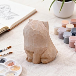 May include: A carved wooden cat figurine in a natural wood tone sits on a white surface. Paint pots in various pastel colors and paintbrushes are nearby, suggesting an art or craft theme. A sketchbook and a potted plant are also in the background.