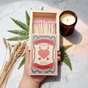 Può includere: Una scatola di fiammiferi in legno con fiammiferi con la punta rosa e un'etichetta decorativa con la scritta "THE PERFECT SINCE KC 2024 MATCH". È visibile anche una candela accesa in un barattolo di vetro marrone.