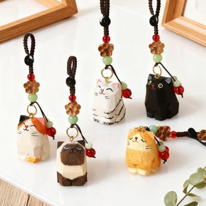 May include: A collection of carved wooden cat figurines in various colors, including white, black, brown, and orange. Each cat has a smiling face and is attached to a brown cord with decorative beads and flowers. The figurines are displayed on a white surface.