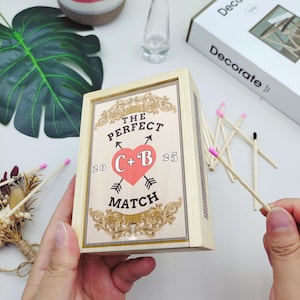 May include: A wooden matchbox with the text "THE PERFECT MATCH" and a heart with the initials "C+B" and the numbers "20" and "25". The match heads are pink. The box is held by a person.