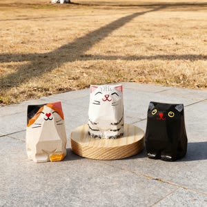 May include: Three wooden cat figurines. One is black, one is white with black stripes, and the third is natural wood with orange and black accents. The cats have smiling faces and are standing on a stone surface.