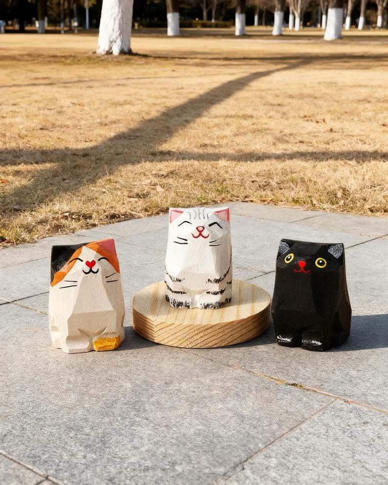 May include: Three wooden cat figurines. One is black, one is white with black stripes, and the third is natural wood with orange and black accents. The cats have smiling faces and are standing on a stone surface.