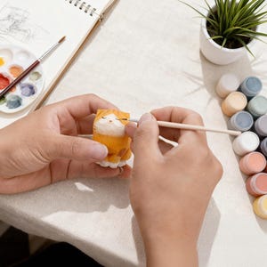 May include: A person is painting a small, orange wooden cat figurine with a paintbrush. The figurine is on a white surface, with a palette of paints, a small potted plant, and a sketchbook nearby. The cat has closed eyes and white paws.
