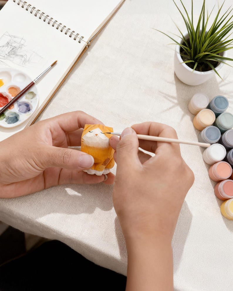 May include: A person is painting a small, orange wooden cat figurine with a paintbrush. The figurine is on a white surface, with a palette of paints, a small potted plant, and a sketchbook nearby. The cat has closed eyes and white paws.