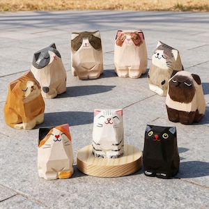 May include: A collection of nine carved wooden cat figurines in various colors and patterns. The cats have smiling faces and are arranged on a gray stone surface. The cats are brown, black, gray, and white.