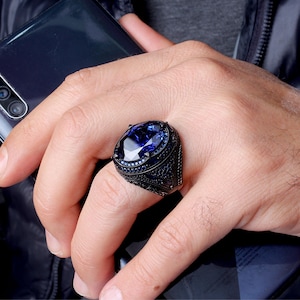May include: A black ring with a large, oval-shaped blue gemstone surrounded by smaller blue gemstones. The ring has intricate details and a black finish.
