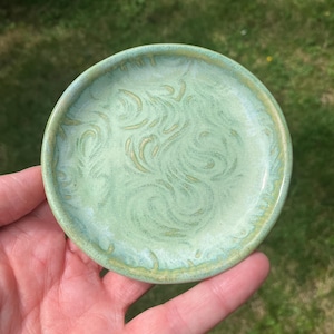 May include: A small, round ceramic dish featuring a light green glaze with a subtle, swirling, textured pattern across the surface. The edges of the dish are slightly raised, and the glaze shows variations in tone with hints of yellow-green.
