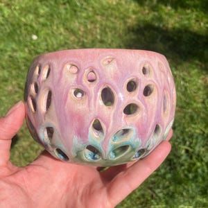 May include: A decorative ceramic bowl with a pierced design. The bowl is glazed in shades of pink, purple, and teal. The holes create a textured surface. The bowl is held in a hand, showing its size and shape. A unique piece for home decor.