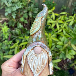 May include: A ceramic gnome figurine featuring a tall, pointed green hat adorned with a sculpted leaf detail. The gnome has a smooth, round pink nose and a textured white beard that tapers to a point. The base is a natural, unglazed tan colour.