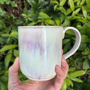 May include: A ceramic mug with a glossy finish featuring a pale cream base color and soft, blended streaks of lavender and pink. The mug has a smooth, rounded body and a curved handle on the right side, showcasing a subtle, ethereal color gradient.