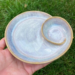 Puede incluir: Un plato redondo de cerámica con un esmalte suave y brillante en tonos azul pálido, lavanda y blanco. El plato incluye una sección circular más pequeña integrada en un lado, con un divisor curvo elevado que separa ambas áreas.