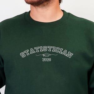 May include: A dark green sweatshirt with the word "STATISTICIAN" arched across the chest in a white, stylized font, with "2026" below. The sweatshirt is a solid color and has long sleeves.