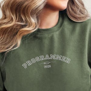 Op de afbeelding: Een olijfgroene sweatshirt met het woord "PROGRAMMER" in witte letters over de borst, met "2026" eronder. De sweatshirt heeft een ronde hals en lange mouwen.