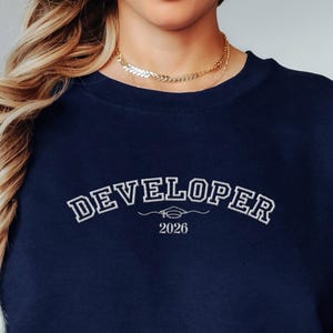 Op de afbeelding: Marineblauwe sweatshirt met het woord "DEVELOPER" in wit geborduurd, gebogen over de borst. Daaronder een afstudeerpet-afbeelding en het jaartal "2026". De sweatshirt is effen van kleur en heeft een ronde hals.