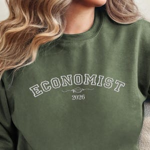 以下が含まれることがあります： オリーブグリーンのスウェットシャツ。白いアーチ状の文字で「ECONOMIST」と書かれています。その下に「2026」と年号が書かれています。クルーネックのスタイルで、柔らかい素材で作られています。