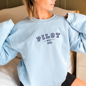 May include: A light blue sweatshirt with the word "PILOT" embroidered in navy blue across the chest. Below the word "PILOT" is a small graphic and the year "2026". The sweatshirt has long sleeves and a crew neck.