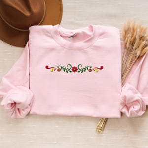 May include: A light pink sweatshirt with a crew neck and long sleeves. The sweatshirt features an embroidered floral design in red, green, and yellow across the chest. A brown hat and dried flowers are in the background.