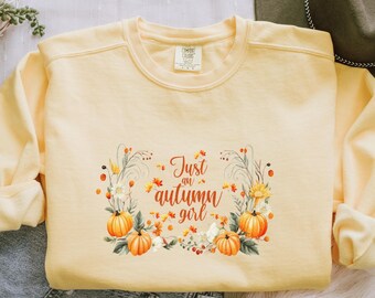 May include: A light yellow sweatshirt with the text "Just an autumn girl" surrounded by a floral wreath with pumpkins and fall foliage. The sweatshirt is made of a soft material.