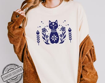 May include: A white t-shirt with a blue cat design. The cat is surrounded by flowers and stars. The text "Comfort Colors" is on the bottom left corner of the shirt.