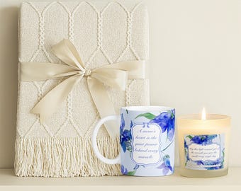May include: A gift set featuring a cream-colored knitted throw blanket tied with a ribbon, a floral-patterned mug with text, and a lit candle in a glass jar. The mug and candle have blue floral designs.
