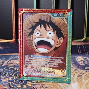 Könnte beinhalten: Eine One Piece Sammelkarte mit Monkey D. Luffy mit Strohhut, eingefasst in einen rechteckigen Rahmen mit roter linker und türkiser rechter Seite, verziert mit goldenen Glitzerkanten. Die Karte zeigt Text und verschiedene Spielstatistiken.