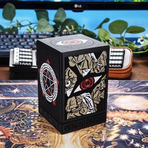 Könnte beinhalten: Schwarze, würfelförmige Kartenaufbewahrungsbox mit Illustrationen im Anime-Stil und einem roten Augendesign. Die Box hat ein Pentagramm-Symbol und Text. Die Box befindet sich auf einer gemusterten Oberfläche.