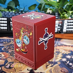Könnte beinhalten: Eine rote, rechteckige Deck-Box mit glitzerndem Finish. Die Vorderseite zeigt eine Grafik einer blaugesichtigen Figur mit Flammen und Perlen, während die Seite ein weißes Totenkopf-Emblem mit gekreuzten Knochen aufweist.