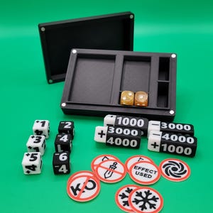 Könnte beinhalten: Ein schwarzer Spiel-Organizer mit Würfeln, Spielsteinen und Zählern auf einer grünen Oberfläche. Das Set enthält weiße Würfel mit schwarzen Zahlen, schwarz-weiße Zähler mit Punktwerten und runde Spielsteine mit verschiedenen Symbolen und Text.
