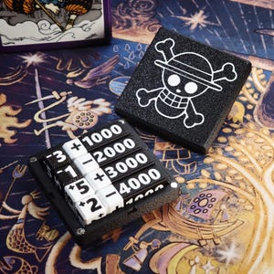 Könnte beinhalten: Schwarz-weißes Spielzubehör zum Thema One Piece. Ein schwarzer Punktezähler mit weißen Zahlen und ein schwarzes Quadrat mit weißem Jolly-Roger-Logo. Die Artikel befinden sich auf einer farbenfrohen, gemusterten Oberfläche.