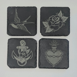 May include: Four square slate coasters with engraved designs. Each coaster features a different design: a swallow, a rose, an anchor with a rose, and a heart with a crown of thorns. The coasters are dark grey.