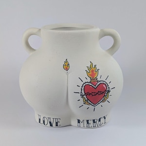 May include: White ceramic vase with two handles, featuring a stylized heart design with flames and the words "LOVE MERCY". The vase has a unique shape and a matte finish, suitable for home decor.