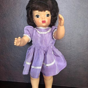 May include: Vintage doll with brown hair styled in a bouffant. The doll wears a lavender dress with white lace trim. The doll has painted facial features, including red lips and large eyes. The doll's arms are outstretched, and it is standing.