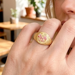 May include: Gold signet ring with a pink bow design. The ring has a textured band and a detailed oval face with a pink bow and floral accents. The ring is a classic piece of jewelry.